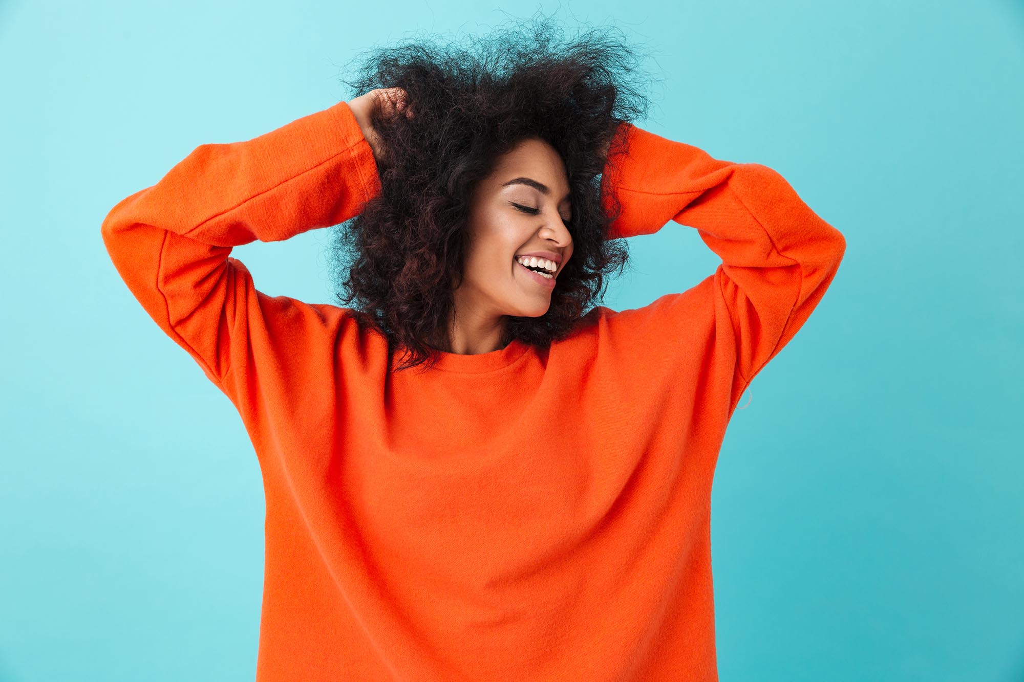 Colorful portrait of smiling woman in orange shirt looking aside and touching her dark curly hair on blue background