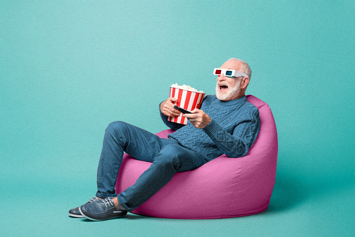 Older man sitting in bean bag chair excitedly watching television while holding a remote, popcorn, and wearing 3d glasses. The background is teal.