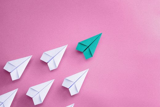 A group of white paper airplanes on a pink surface. They are uniform, except the leading paper airplane is a teal color, and all are pointed the same direction.