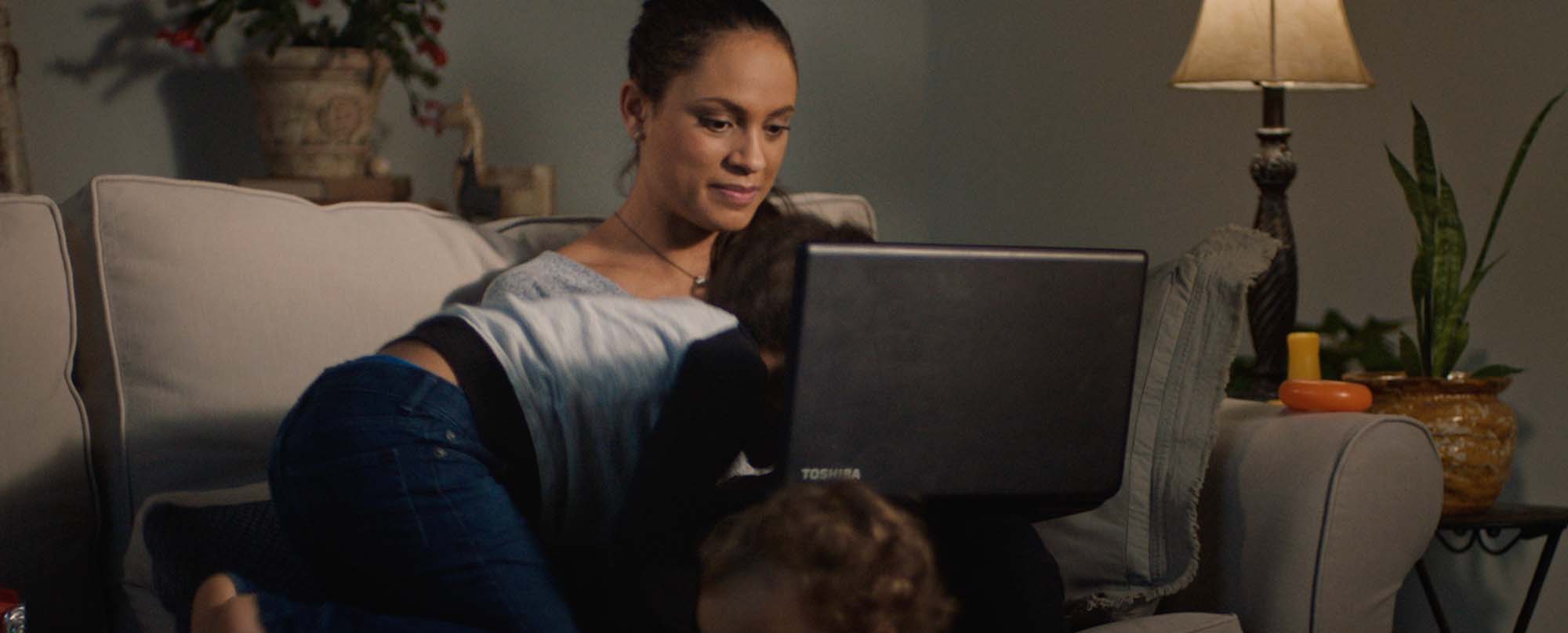 Woman sitting on couch using laptop to bank while children run by and climb on top of her.