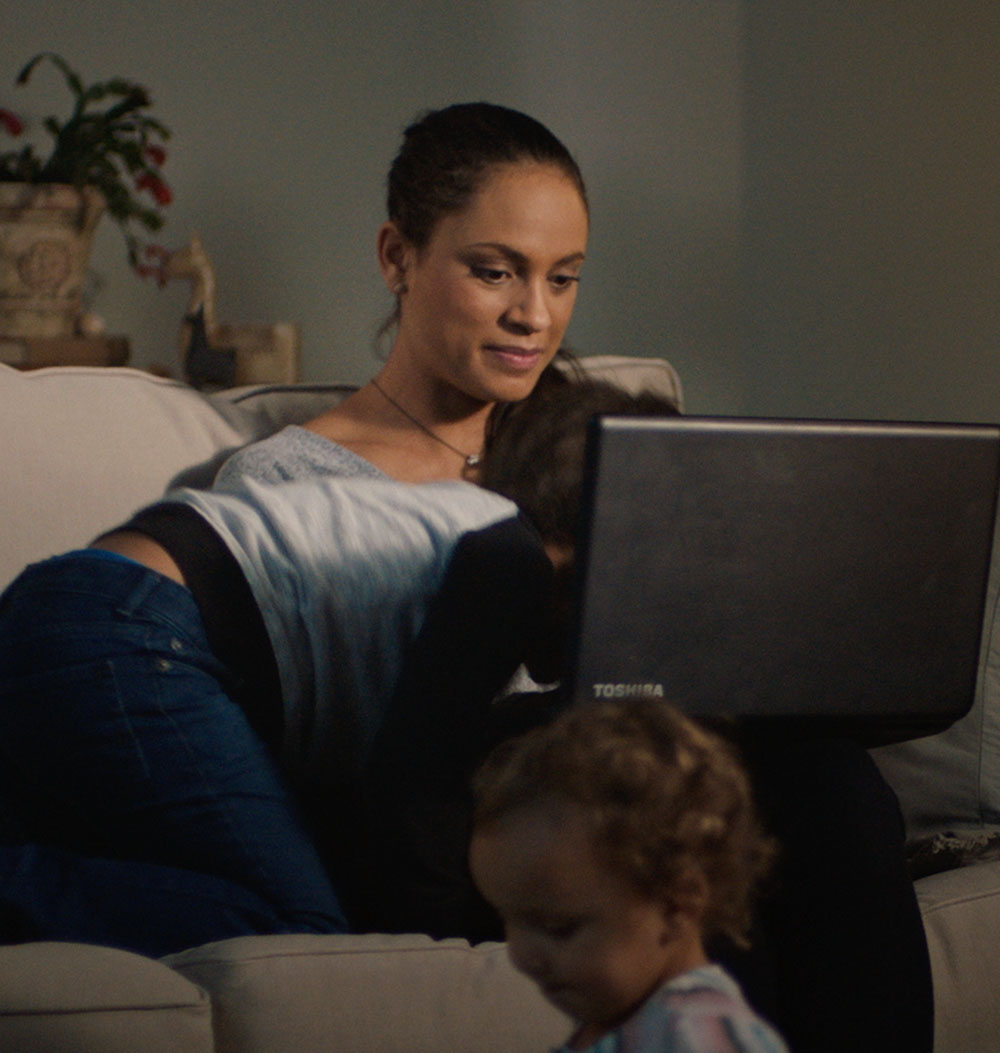 Woman sitting on couch using laptop to bank while children run by and climb on top of her.