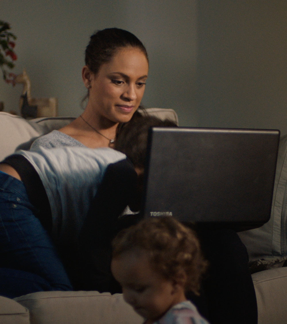 Woman sitting on couch using laptop to bank while children run by and climb on top of her.