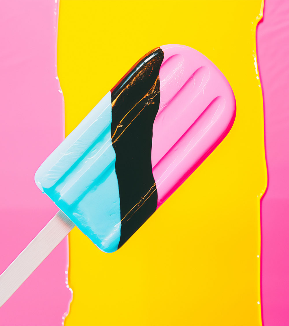 A blue, black and pink popsicle on a white stick against yellow and pink background. Minimalist and abstract feel.