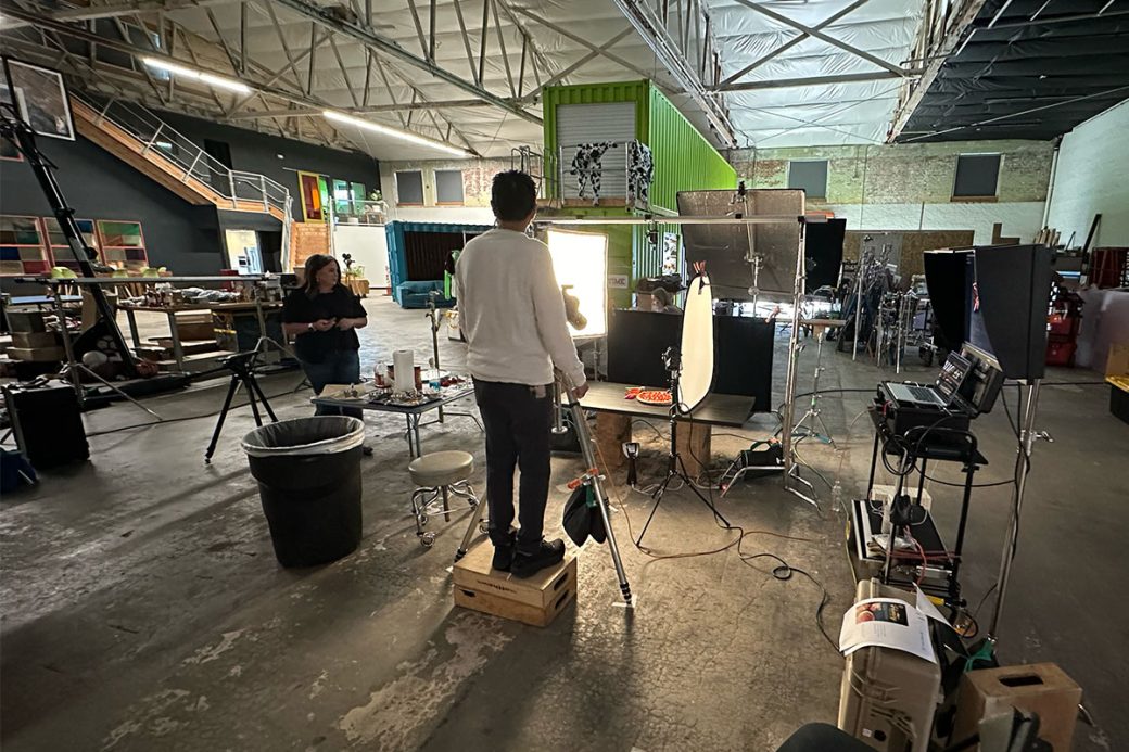 A behind the scenes photograph of a food photoshoot. There is a photographer looking through his camera at a pizza that is surrounded by photo equipment. The photoshoot is taking place in a large industrial warehouse space.