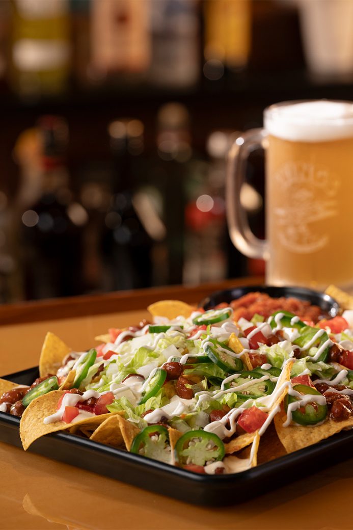 A tray of nachos is topped with beans, diced tomatoes, thinly sliced jalapeños, shredded lettuce and drizzled with sour cream. One of the tortilla chips is hanging out of the tray, and there is a drink out of focus in the background.