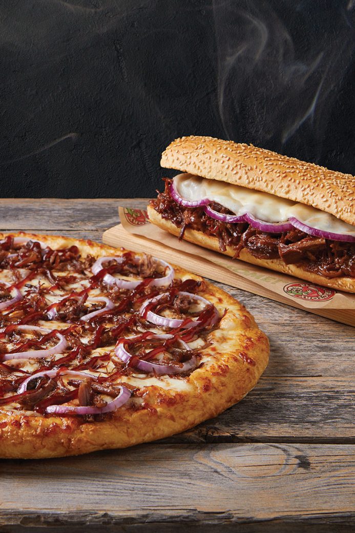 A promotional photo showing a bbq brisket pizza next to a bbq brisket sandwich. There is steam rising from the sandwich. Both items sit on top of a rugged, wooden surface.