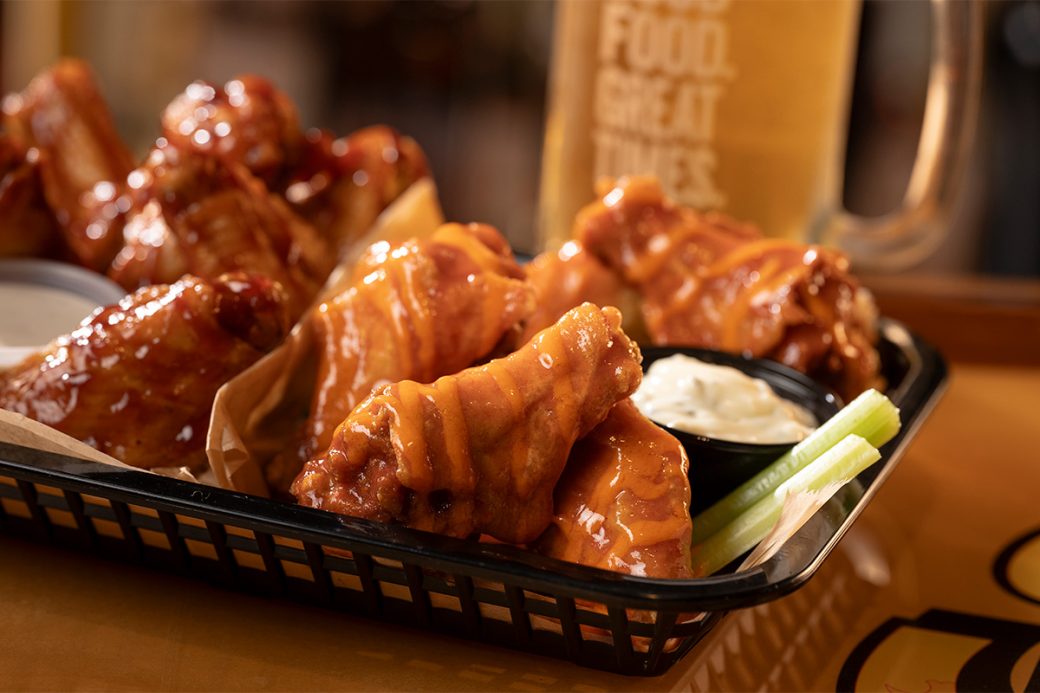 A promotional photo of a basket of chicken wings. The wings are covered in two different kinds of hot sauce. There are also two sticks of celery and a small bowl of blue cheese in the basket with the wings. There is a drink out of focus in the background.