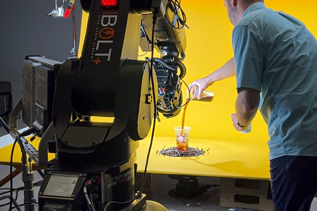 A behind the scenes image of a robot arm filming a man pouring cold coffee into a Scooter's Coffee cup. The cup is surrounded by coffee beans, and sits on top of a solid yellow backdrop.