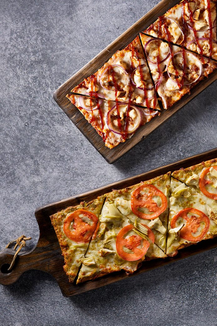 Two flatbreads are shown from overhead, and at angles next to each other. Both sit on wooden paddles. The top flatbread has cheese, sliced chicken, cooked red onions and a drizzle of bbq sauce. The other flatbread is topped with a pesto sauce, cheese, sliced tomatoes an artichokes.