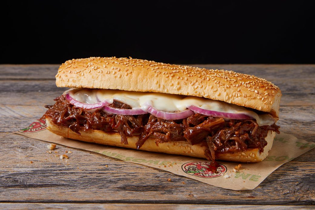 A promotional photo showing a bbq brisket sandwich. There is steam rising from the sandwich and it sits on top of a rugged, wooden surface.