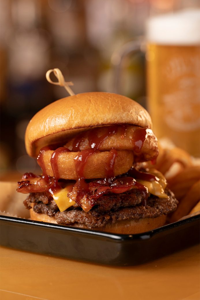 A burger drizzled with bbq sauce, and stacked with two onion rings, crispy slices of bacon and a slice of cheddar cheese sits in a black metal tray. There is a beer that is out of focus in the background.