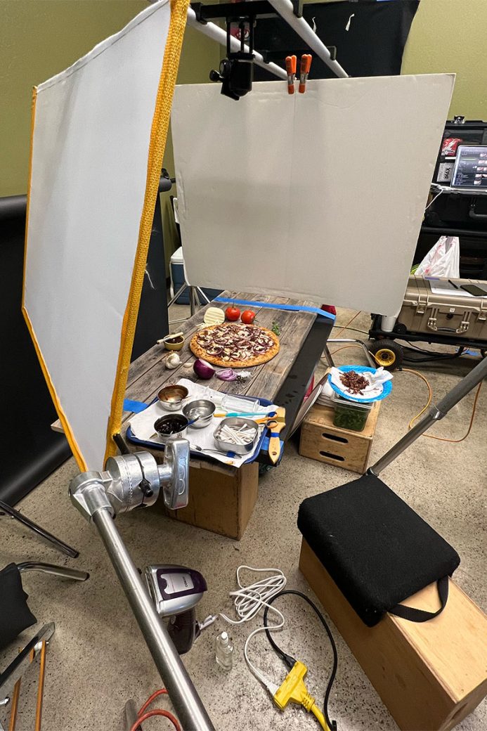 A behind the scenes photo of a food photoshoot. There are large white boards above a pizza, and various styling supplies and photo equipment can be seen around the room.