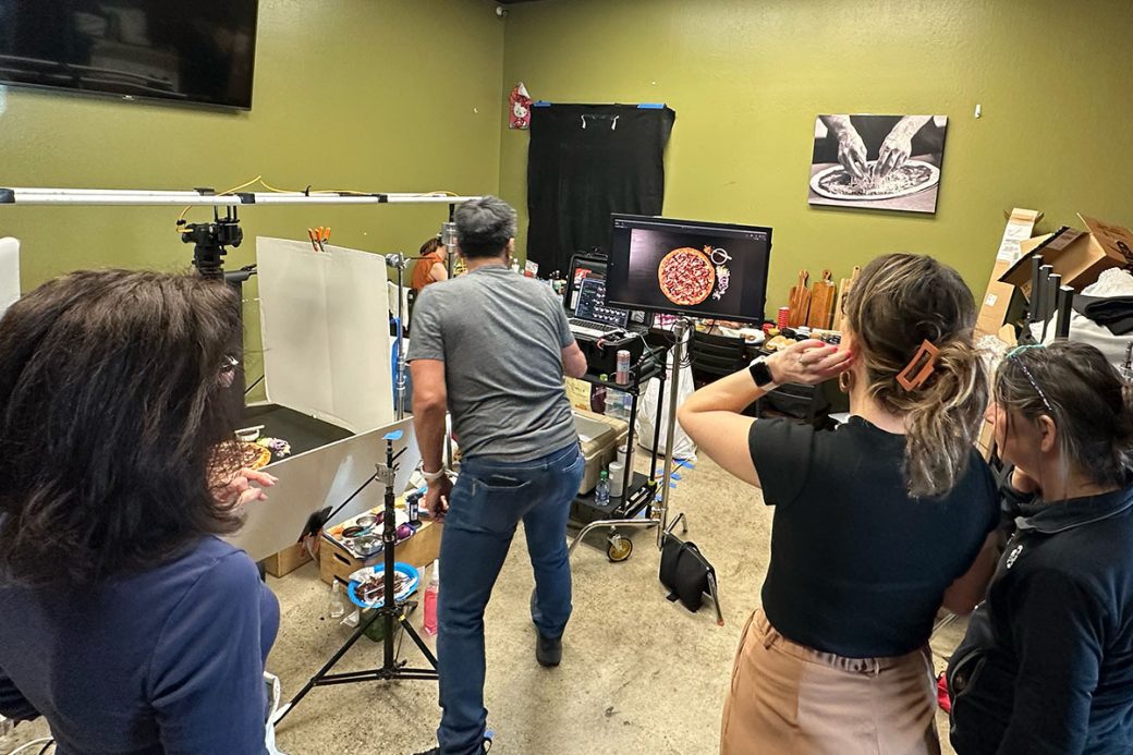 A behind the scenes photograph of a food photoshoot. There is a photographer looking at a monitor, while a food stylist, creative director, and client look on and direct. There is photo and styling equipment in the background.