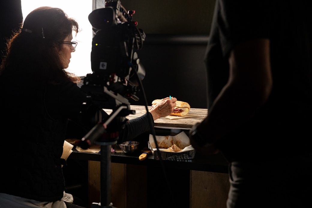 A food stylist carefully places an ingredient on a BBQ Brisket Sandwich on a photo set. There is a large light and a camera on either side of her.