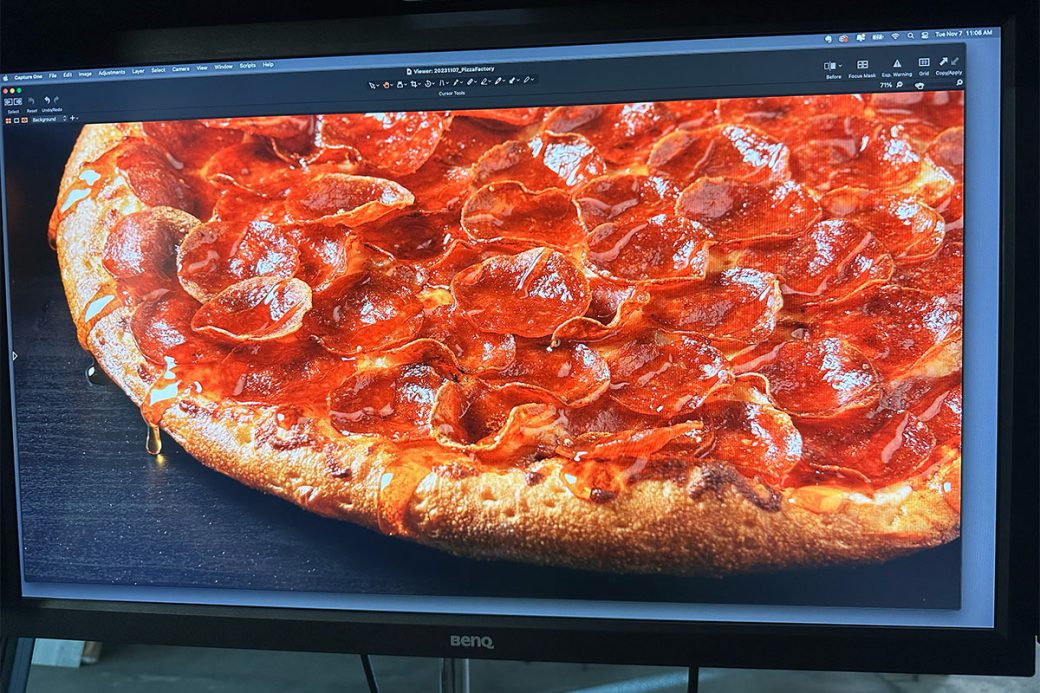 A monitor at a photoshoot displays a pepperoni pizza with hot honey drizzle dripping off of the crust. The pizza is sitting on a dark wood surface.