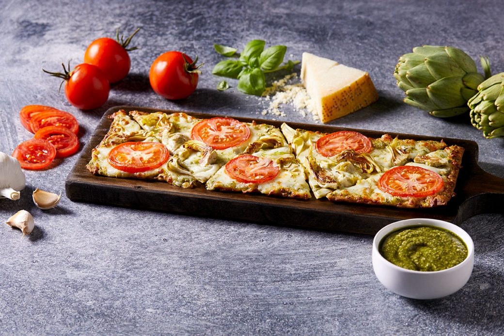 A pesto artichoke flatbread sits on a wooden paddle. It sits next to garlic cloves, tomatoes, basil, a block of parmesan cheese, artichokes and a bowl of pesto sauce.