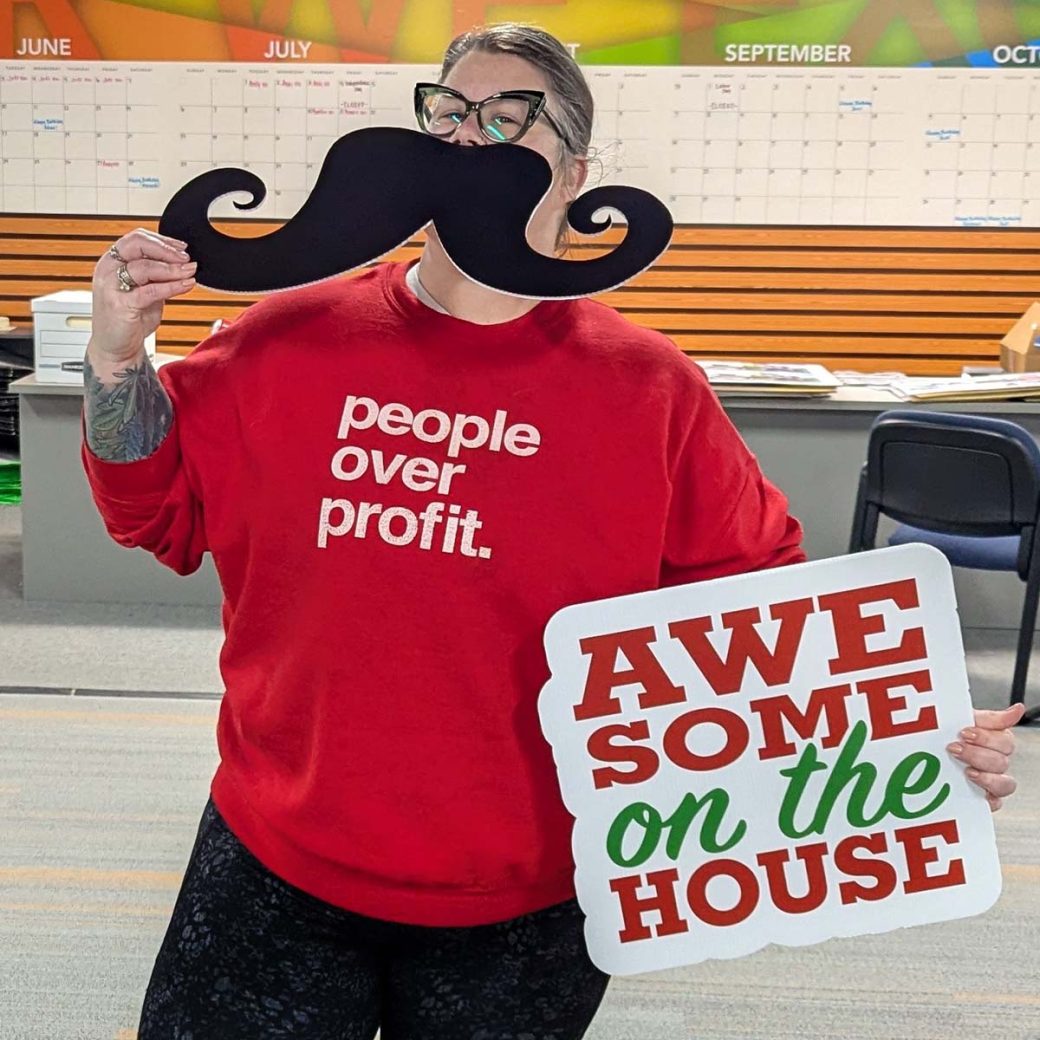 Kirsten Hamrick Hinkson holding an oversized mustache over her mouth and another sign that says "awesome on the house" in her other hand