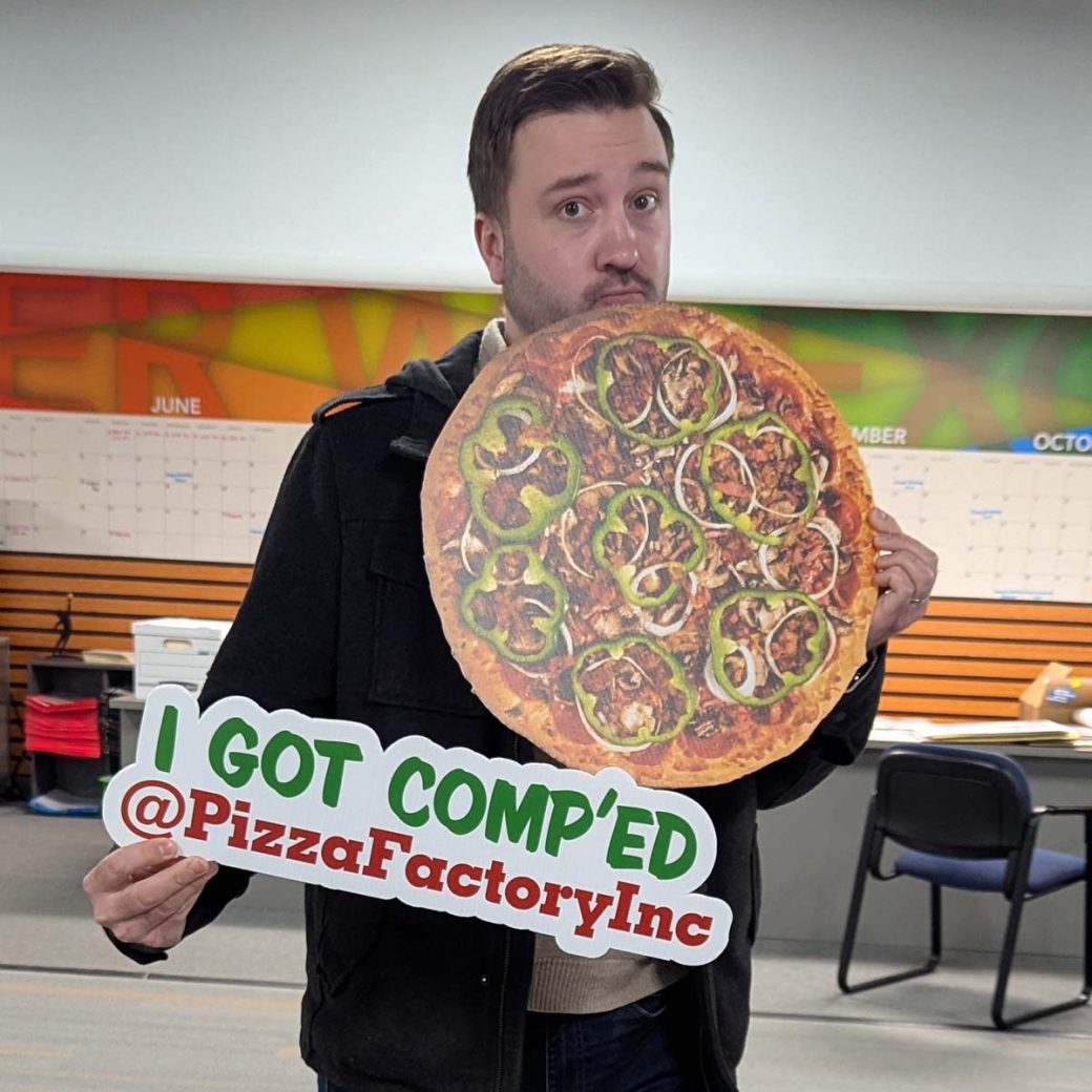 Sean O'Leary, Asher Senior Art Director, poses with photo props for Pizza Factory. There is a large pizza in one of his hands, and in the other is a sign that reads "I got comp'ed @PizzaFactoryInc"