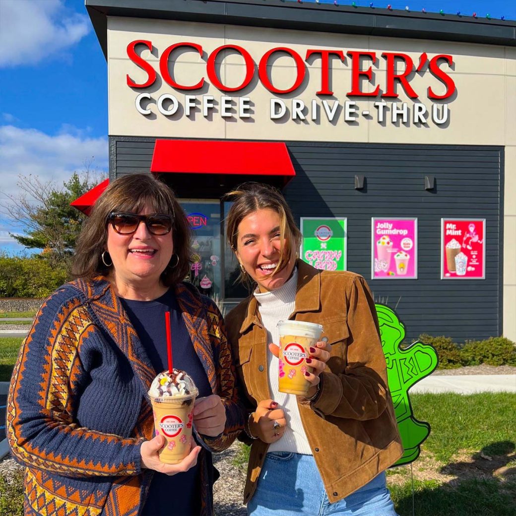 Marina McQueary and Alice Talarico smiling together and holding iced coffee drinks in front of a Scooter's Coffee Drive-Thru